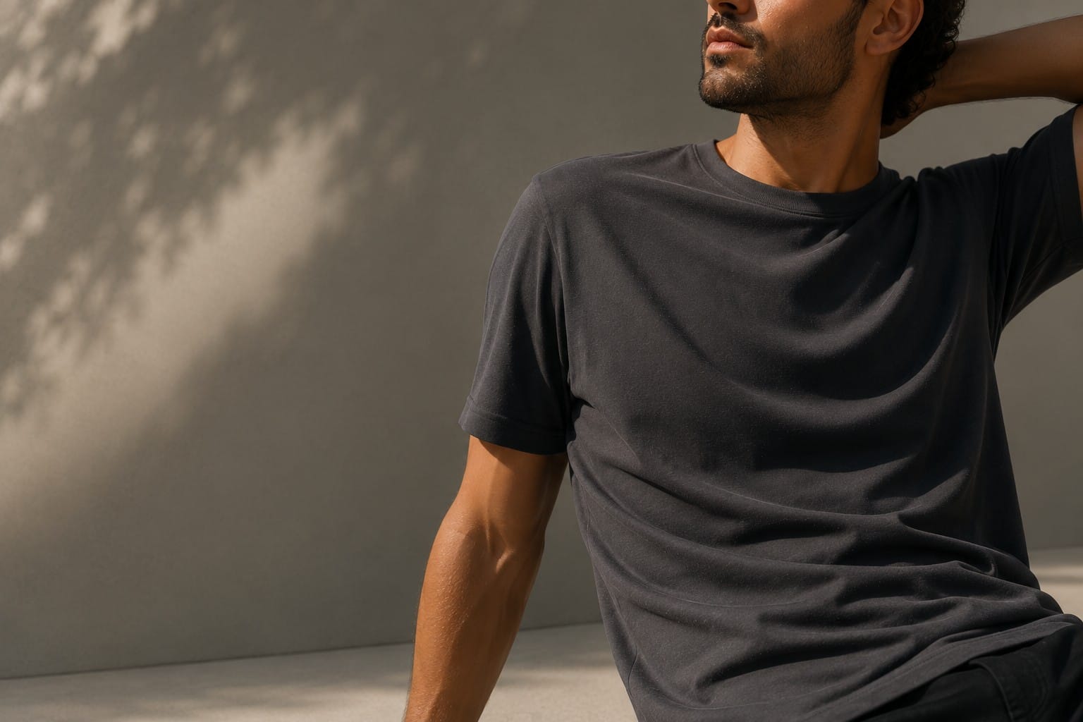 A person wearing a soft charcoal Fibre52 Verified t-shirt, seated in dappled natural light against a textured wall.